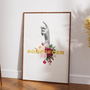 May include: A framed art print featuring a grayscale hand pointing upwards, adorned with a bouquet of flowers in shades of pink, red, and cream. The gold text "AGE DECER" is below the hand. The artwork is displayed against a white wall.