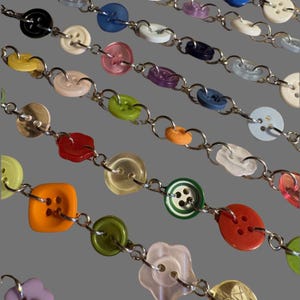 Puede incluir: Primer plano de varias hebras de joyería de botones. Los botones son de varios colores, incluyendo negro, blanco, rosa, verde y naranja. Están conectados por eslabones de cadena plateados. Los botones tienen diferentes formas y tamaños.