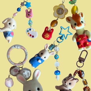 May include: A collection of five keychains featuring flocked animal figurines, including a white puppy, a fawn, a rabbit, and a squirrel. The keychains are decorated with colorful plastic beads, charms shaped like stars, flowers, mushrooms, and a paint palette.
