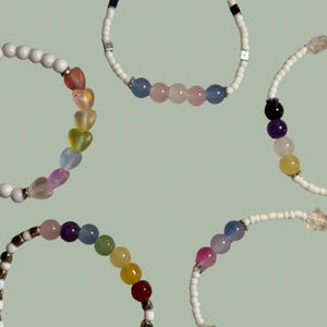 Puede incluir: Varias collares de cuentas con diversos diseños. Un collar presenta cuentas en forma de corazón en colores del arcoíris. Otros collares tienen cuentas redondas en tonos de rosa, azul y otros colores. Sobre un fondo verde claro.