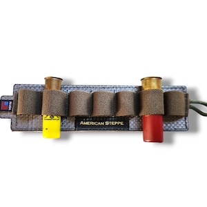 May include: A grey and brown shotgun shell holder with six loops. The holder has the text "AMERICAN STEPPE" and a small American flag patch. Two shotgun shells are visible, one yellow and one red.