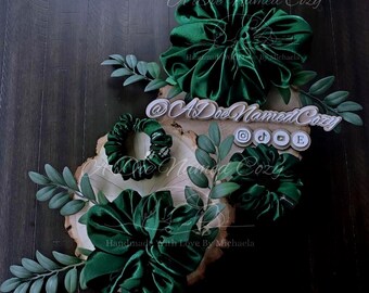 May include: An arrangement of green velvet scrunchies and floral accents on a wooden slice. The scrunchies are in various sizes and styles. The text "A Doe Named Cozy" and social media icons are visible.