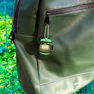 May include: An olive green backpack with a smooth finish and a black zipper, featuring a small digital watch keychain attached to the zipper pull. The watch has a black face with a bright green border and displays the time 4:15 and date WED 4-5.