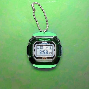 May include: A digital keychain watch with a translucent dark green square case and a bright green top and bottom bezel. The watch features a silver ball chain, a digital display showing the time 3:58, the date 4-5, and the text SKMEI LUMINESCENT.