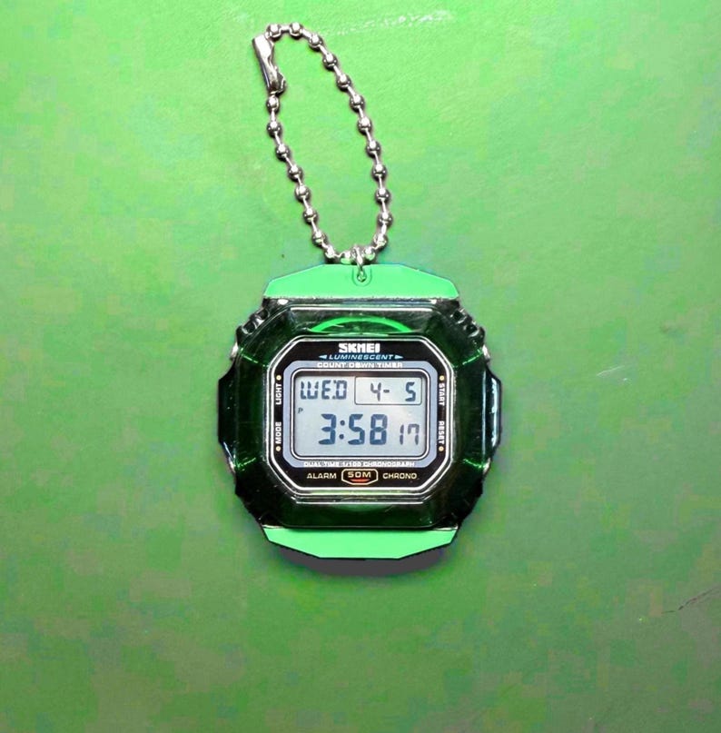 May include: A digital keychain watch with a translucent dark green square case and a bright green top and bottom bezel. The watch features a silver ball chain, a digital display showing the time 3:58, the date 4-5, and the text SKMEI LUMINESCENT.