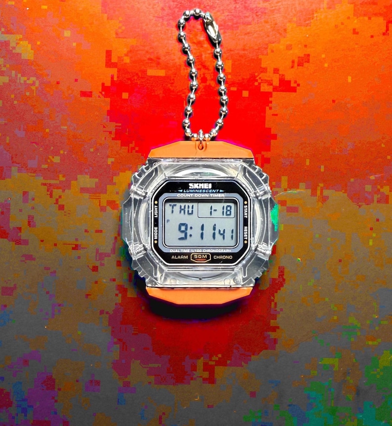 May include: A digital stopwatch with a clear, transparent plastic casing and orange accents. The face displays the text SKMEI, LUMINESCENT, COUNT DOWN TIMER, and shows the time 9:11:41 on Thursday, January 18. It includes a silver metal ball chain.