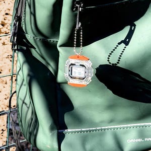 May include: A small digital watch keychain featuring a clear, transparent casing with bright orange accents on the top and bottom. The watch face displays a digital time and date, and it is attached to a silver-toned metal ball chain.