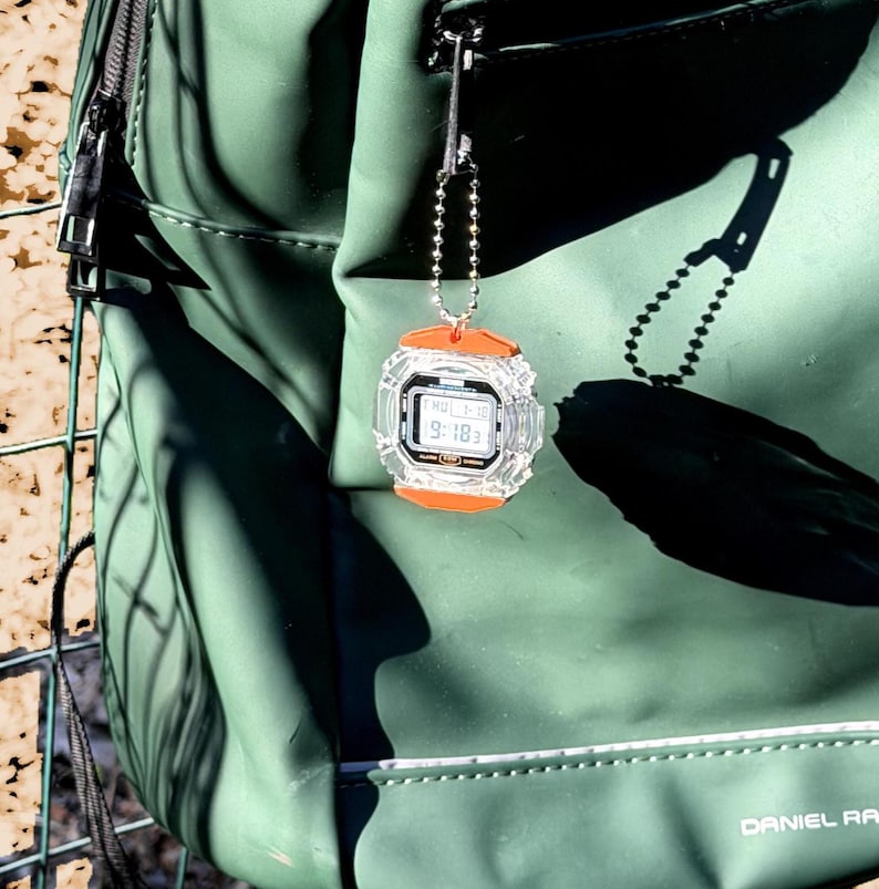 May include: A small digital watch keychain featuring a clear, transparent casing with bright orange accents on the top and bottom. The watch face displays a digital time and date, and it is attached to a silver-toned metal ball chain.
