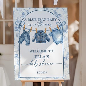 以下が含まれることがあります： 木製のイーゼルには、青と白の花柄のデザインのベビーシャワーのサインが表示されています。「A Blue Jean Baby is on the Way」と書かれており、8.2.2025にEllaのベビーシャワーにゲストを歓迎します。サインにはベビー服のイラストが描かれています。