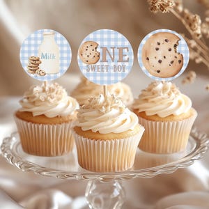 Könnte beinhalten: Cupcakes mit weißem Zuckerguss und dekorativen Aufsätzen auf einer Glas-Kuchenplatte. Die Aufsätze zeigen eine Milchflasche, einen Keks und den Text "ONE SWEET BOY" auf blauem Vichy-Hintergrund. Die Cupcakes sind hellbraun.