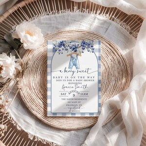 May include: A baby shower invitation with a blue and white gingham pattern. The invitation features the text "a berry sweet" and details for the event, including the date, time, and location. The design includes blueberries and a pair of overalls.