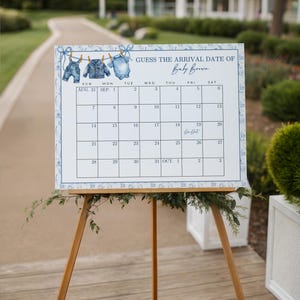 May include: A baby shower calendar on a wooden easel. The calendar has a blue and white floral border and features a clothesline with baby clothes illustrations. The text reads "Guess the Arrival Date of Baby Brown."