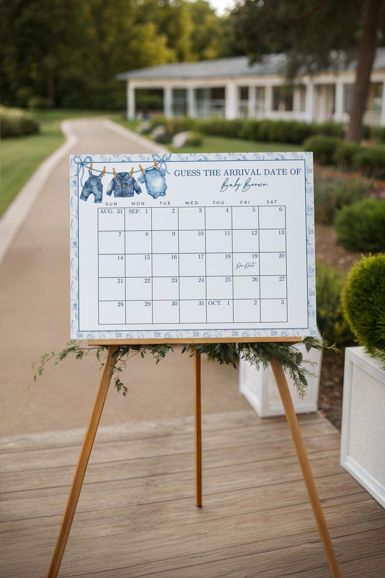 May include: A baby shower calendar on a wooden easel. The calendar has a blue and white floral border and features a clothesline with baby clothes illustrations. The text reads "Guess the Arrival Date of Baby Brown."