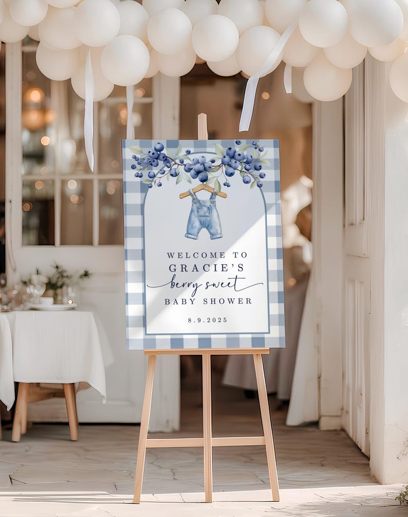May include: A baby shower sign on a wooden easel. The sign has a blue and white gingham border and features the text "Welcome to Gracie's berry sweet baby shower" with a watercolor illustration of blueberries and overalls. White balloons are in the background.