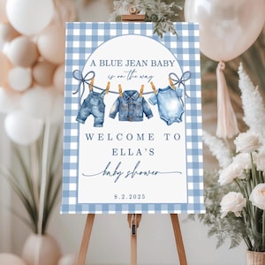 May include: A baby shower sign with a blue gingham background and the text "A BLUE JEAN BABY is on the way." The sign features illustrations of baby clothes hanging on a clothesline. The sign reads "WELCOME TO ELLA'S baby shower 8.2.2025."