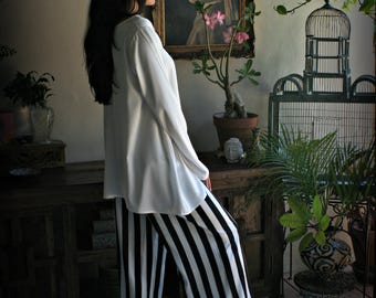 May include: A woman wearing a white blouse and black and white striped pants stands in a room with a painting and plants. The painting is in a gold frame. The pants have wide vertical stripes. The room has a vintage aesthetic.