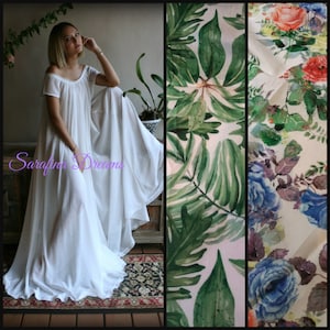 May include: A white maxi dress with a scoop neck and short sleeves. The dress is made from a lightweight fabric and has a flowing silhouette. The dress is shown on a model in a room with a plant in the background. The dress is available in a variety of floral prints, including a tropical leaf print and a floral print with blue roses.