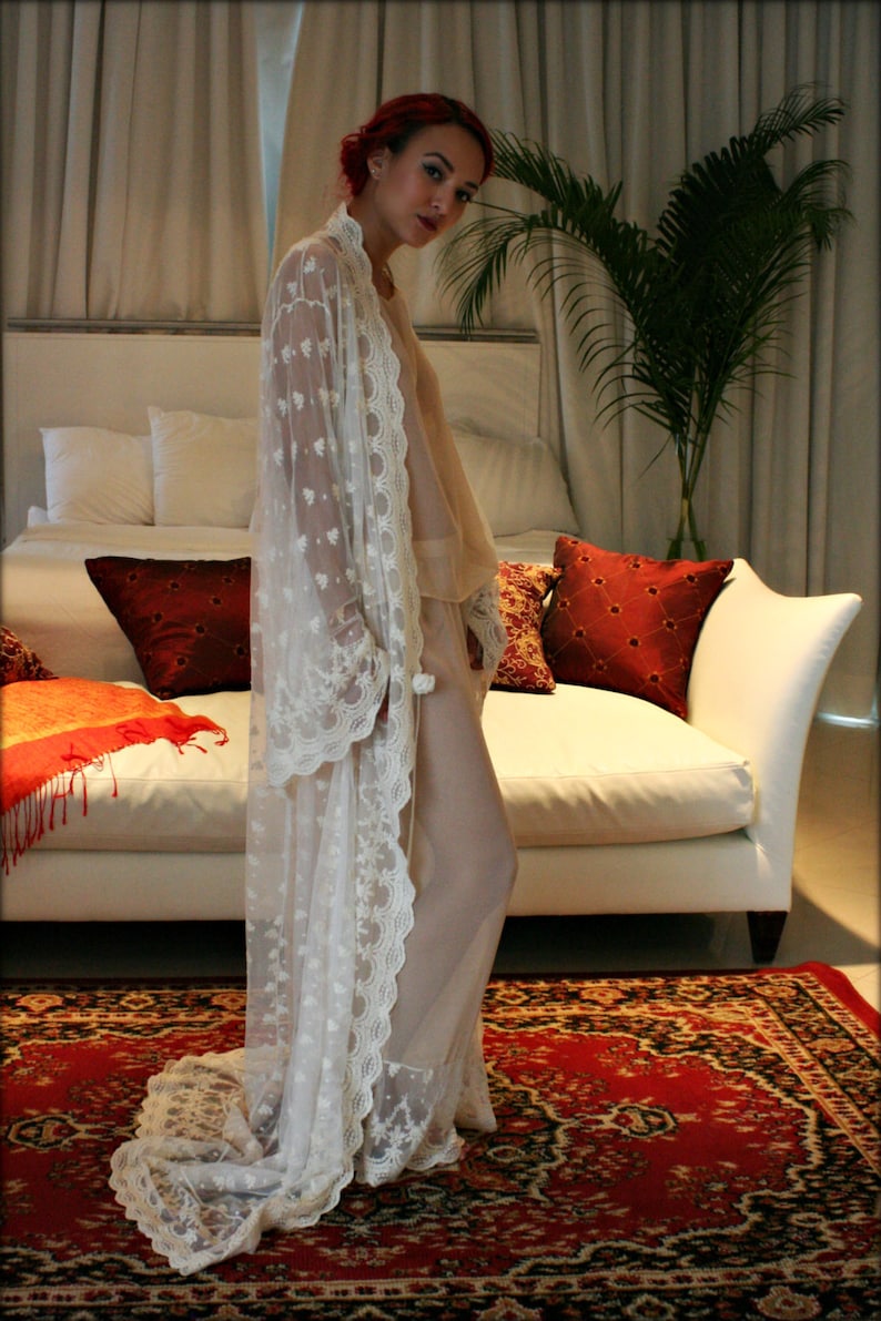 May include: A woman wearing a white lace robe with a sheer overlay. She is standing in a bedroom with a white sofa and a red and black patterned rug.