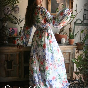 May include: A white maxi dress with a floral print featuring pink, red, blue, and green flowers. The dress has long sleeves with a gathered puff at the shoulder and a cinched waist.