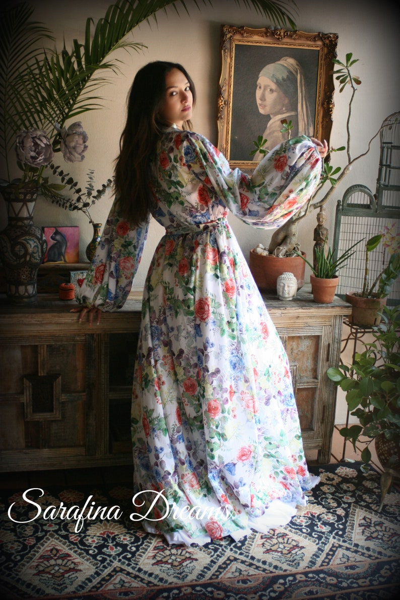 May include: A white maxi dress with a floral print featuring pink, red, blue, and green flowers. The dress has long sleeves with a gathered puff at the shoulder and a cinched waist.