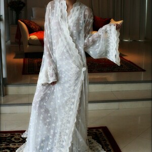 May include: A white lace robe with long sleeves and a wrap closure. The robe is sheer and has a delicate floral pattern. The model is wearing the robe and standing on a patterned rug.