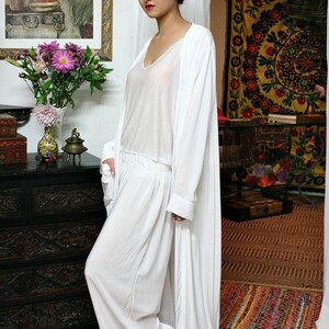 May include: A white three-piece ensemble consisting of a sleeveless V-neck top, wide-leg trousers with an elasticized waistband, and a long-sleeved, floor-length open duster cardigan. The garments are made of a lightweight, semi-sheer white fabric.
