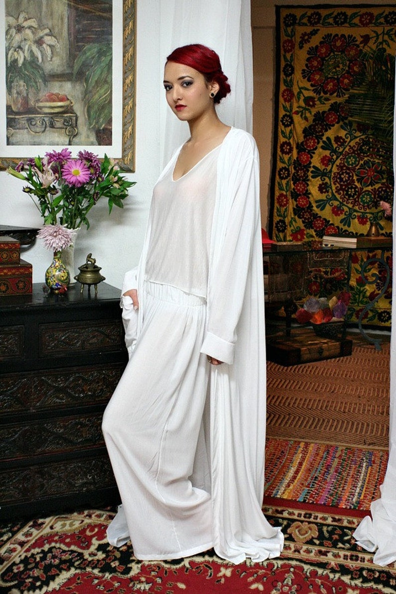 May include: A white three-piece ensemble consisting of a sleeveless V-neck top, wide-leg trousers with an elasticized waistband, and a long-sleeved, floor-length open duster cardigan. The garments are made of a lightweight, semi-sheer white fabric.