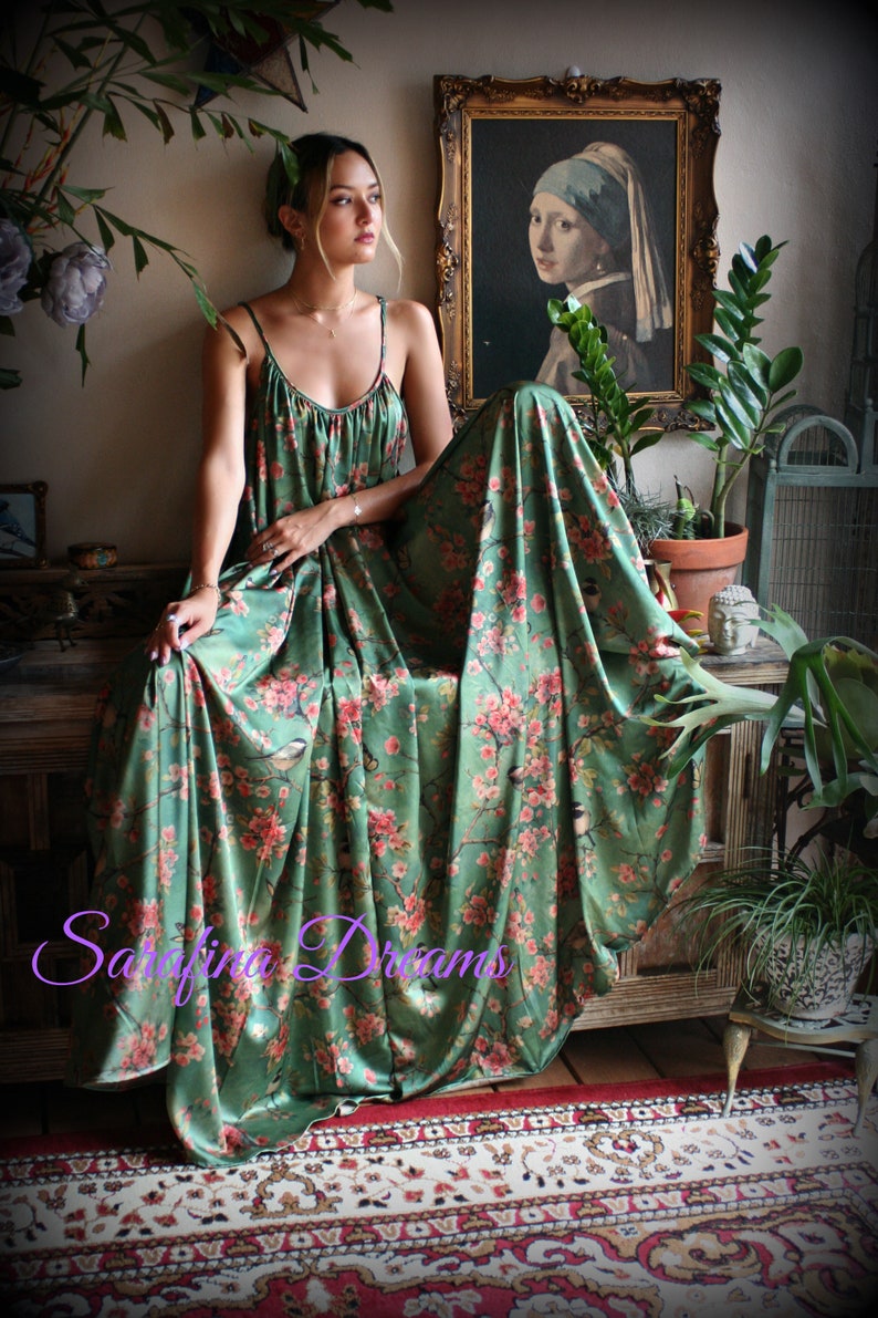 Puede incluir: Un vestido largo verde con un estampado floral de flores rosas y p&aacute;jaros. El vestido tiene tirantes finos y cae elegantemente. El texto "Sarafina Dreams" es visible en la imagen.
