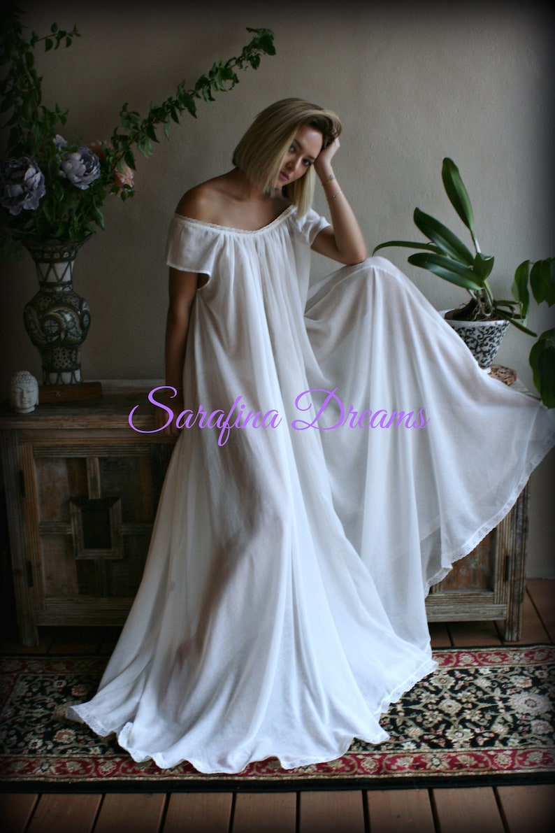May include: A white off-the-shoulder maxi dress with a flowing skirt. The dress is made of a lightweight fabric and has a relaxed fit. The model is sitting on a wooden bench with a floral arrangement and a potted plant in the background. The text "Sarafina Dreams" is visible in the image.