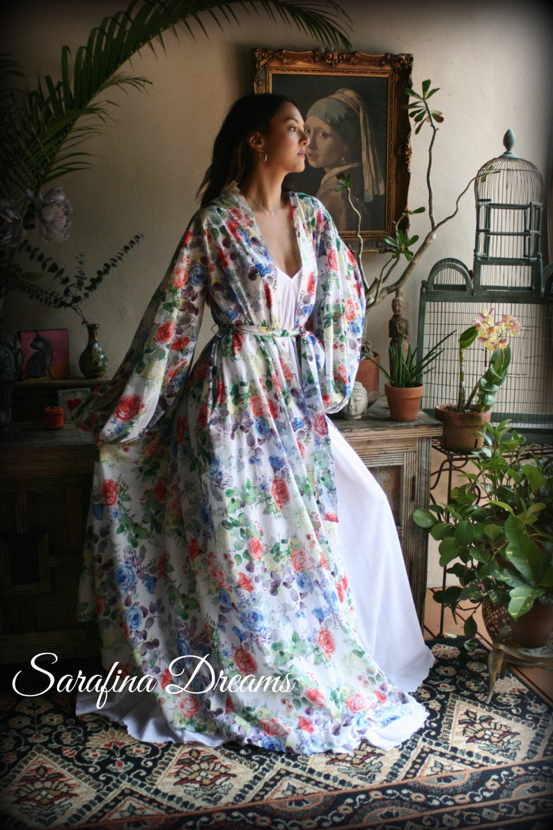 May include: A white floral print kimono with long sleeves and a sash. The kimono is worn over a white slip dress. The model is standing in a room with plants and a painting.