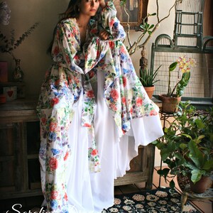 May include: A woman wearing a white and floral print maxi dress with long sleeves. The dress has a flowing skirt and a cinched waist. The woman is standing in a room with a rug, plants, and a birdcage.
