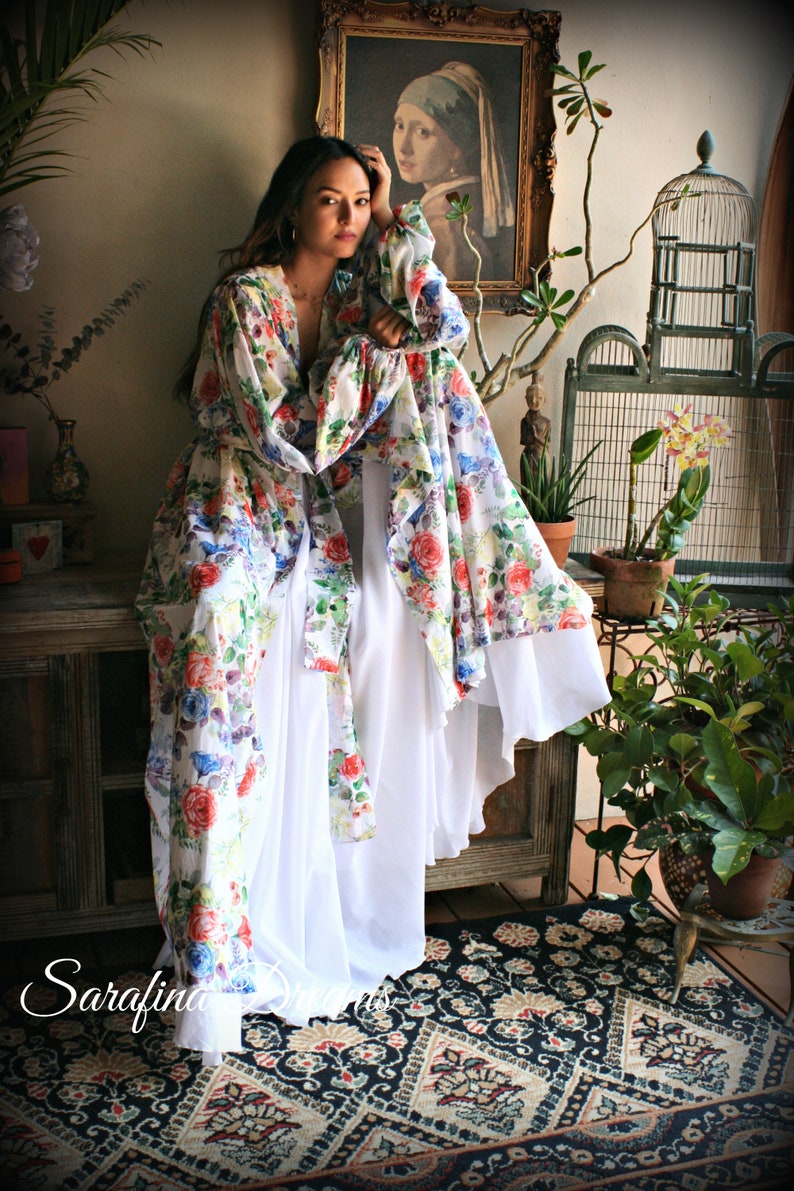 May include: A woman wearing a white and floral print maxi dress with long sleeves. The dress has a flowing skirt and a cinched waist. The woman is standing in a room with a rug, plants, and a birdcage.