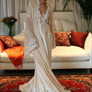 May include: A woman wearing a long, white, lace-trimmed robe with a satin slip underneath. She is standing on a red and black patterned rug in a room with a white couch and pillows.