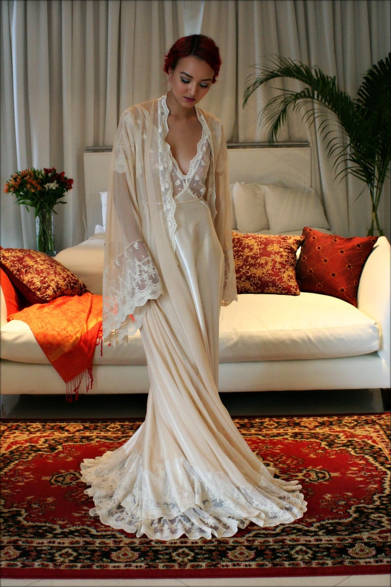 May include: A woman wearing a long, white, lace-trimmed robe with a satin slip underneath. She is standing on a red and black patterned rug in a room with a white couch and pillows.