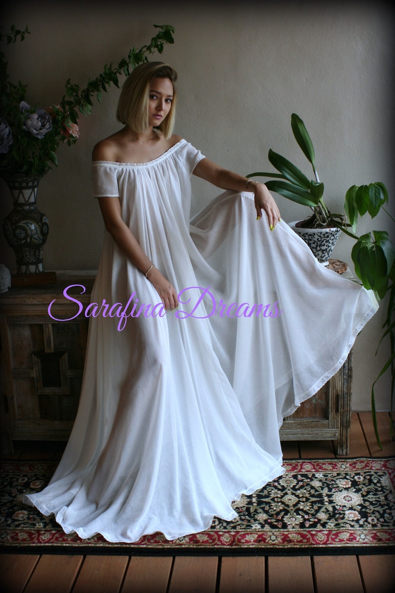 May include: A white off-the-shoulder maxi dress with a gathered neckline and short sleeves. The dress is made of a sheer fabric and has a lace trim at the hem. The dress is being worn by a person in a room with plants and a wooden chest.