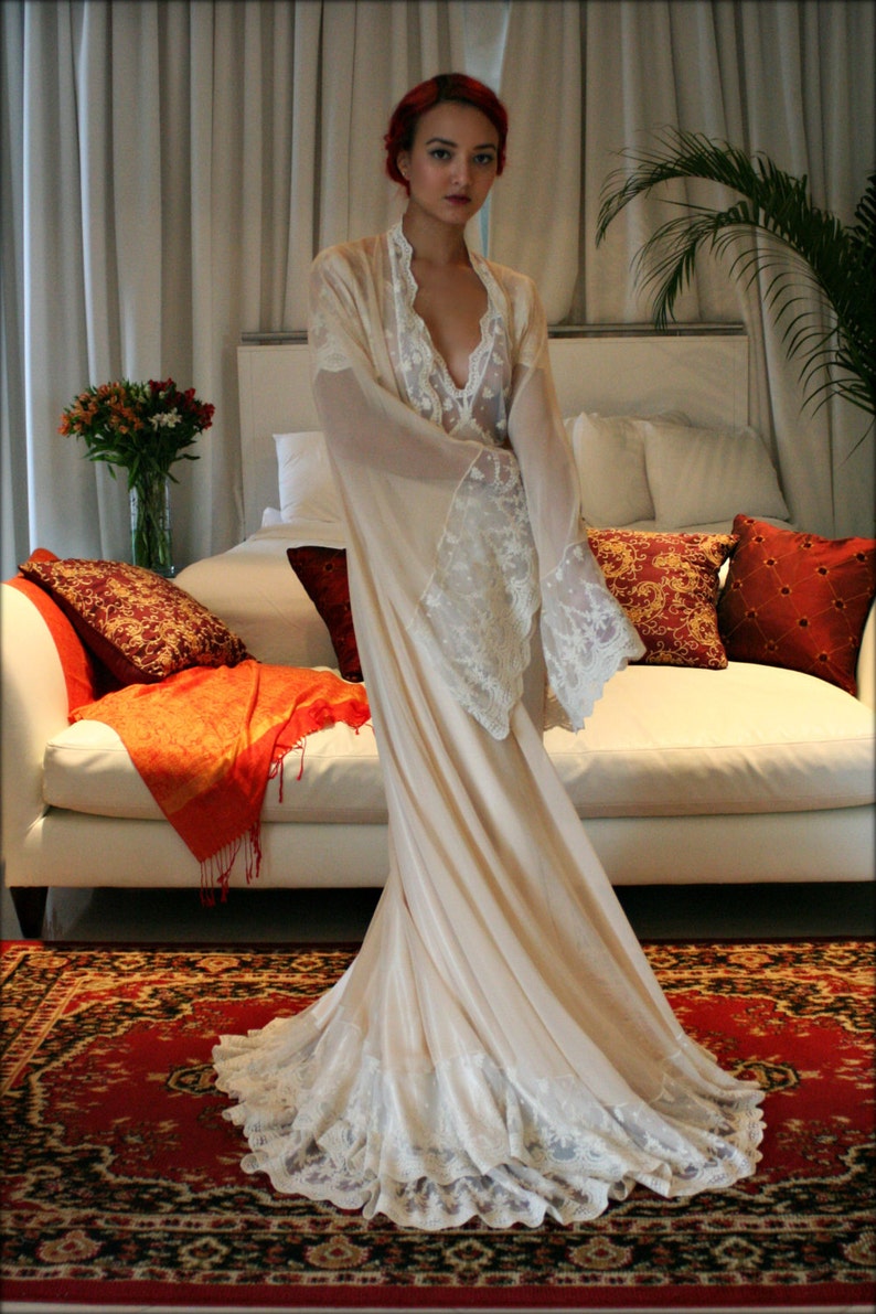 May include: A woman with red hair wearing a white lace robe with a long flowing skirt. She is standing in a room with a white couch and a rug with a floral pattern.