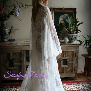 K&ouml;nnte beinhalten: Ein wei&szlig;es Brautkleid aus Spitze mit langen &Auml;rmeln und einem flie&szlig;enden Rock. Das Kleid hat einen tiefen V-Ausschnitt und zarte Spitzendetails. Das Model steht in einem Raum mit einem Holzschrank und einem Teppich.