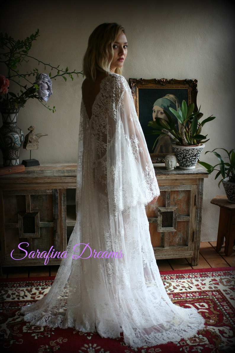 K&ouml;nnte beinhalten: Ein wei&szlig;es Brautkleid aus Spitze mit langen &Auml;rmeln und einem flie&szlig;enden Rock. Das Kleid hat einen tiefen V-Ausschnitt und zarte Spitzendetails. Das Model steht in einem Raum mit einem Holzschrank und einem Teppich.