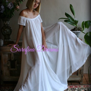 May include: A white off-the-shoulder maxi dress with a flowing skirt and short sleeves. The dress is made of a sheer fabric and has a relaxed fit. The model is standing on a wooden floor with a rug underneath. The text "Sarafina Dreams" is visible in the image.