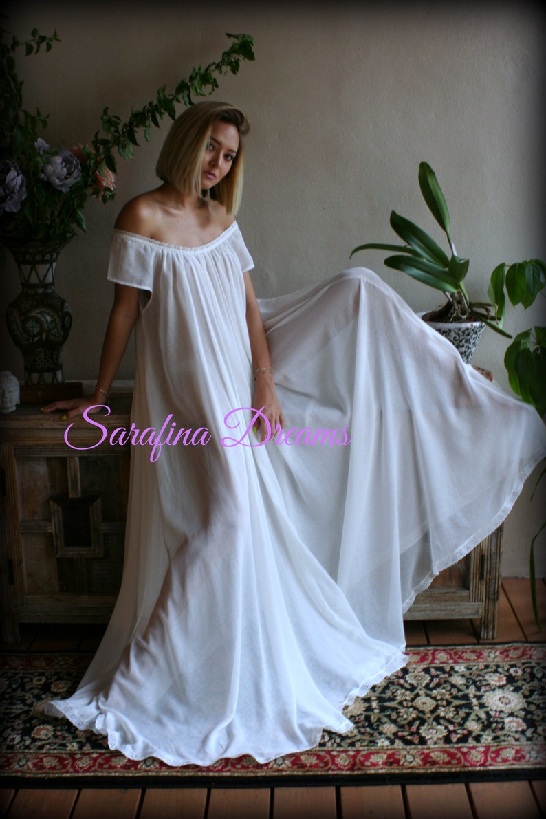May include: A white off-the-shoulder maxi dress with a flowing skirt and short sleeves. The dress is made of a sheer fabric and has a relaxed fit. The model is standing on a wooden floor with a rug underneath. The text "Sarafina Dreams" is visible in the image.