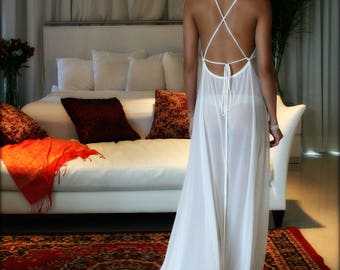 Könnte beinhalten: Ein weißes, durchsichtiges Maxikleid mit einem Kreuzrücken-Detail und einer Spitzenborte. Das Kleid ist fließend und elegant, und das Model steht in einem Schlafzimmer mit einem weißen Sofa und einem gemusterten Teppich.