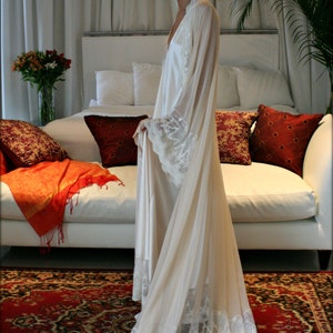 May include: A woman wearing a white satin robe with lace trim, standing in front of a white couch with red and gold pillows. The couch is in a room with white curtains and a patterned rug.