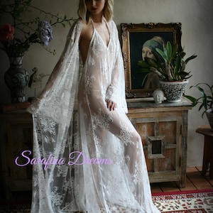 K&ouml;nnte beinhalten: Ein wei&szlig;es Spitzenkleid mit langen &Auml;rmeln und einem tiefen V-Ausschnitt. Das Kleid ist durchsichtig und hat ein zartes Blumenmuster. Das Model tr&auml;gt das Kleid &uuml;ber einem passenden Slip.