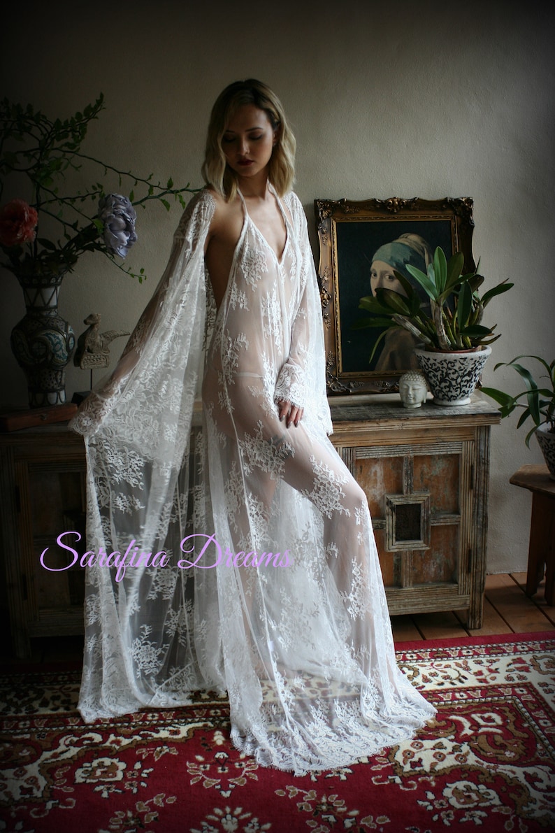 K&ouml;nnte beinhalten: Ein wei&szlig;es Spitzenkleid mit langen &Auml;rmeln und einem tiefen V-Ausschnitt. Das Kleid ist durchsichtig und hat ein zartes Blumenmuster. Das Model tr&auml;gt das Kleid &uuml;ber einem passenden Slip.