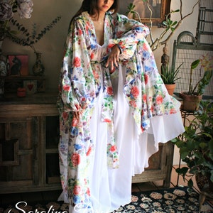 May include: A white floral print robe with long sleeves and a white slip underneath. The robe has a gathered waist and a flowing silhouette. The model is standing on a patterned rug.