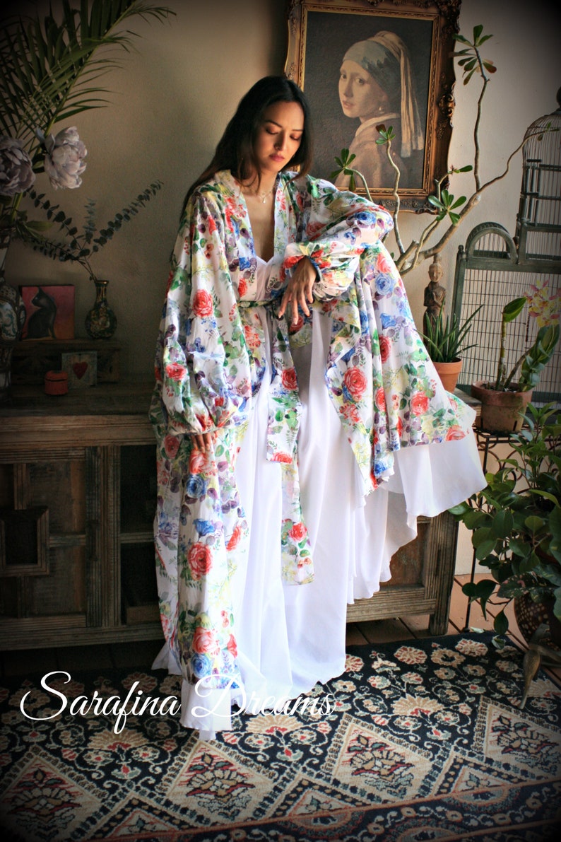 May include: A white floral print robe with long sleeves and a white slip underneath. The robe has a gathered waist and a flowing silhouette. The model is standing on a patterned rug.