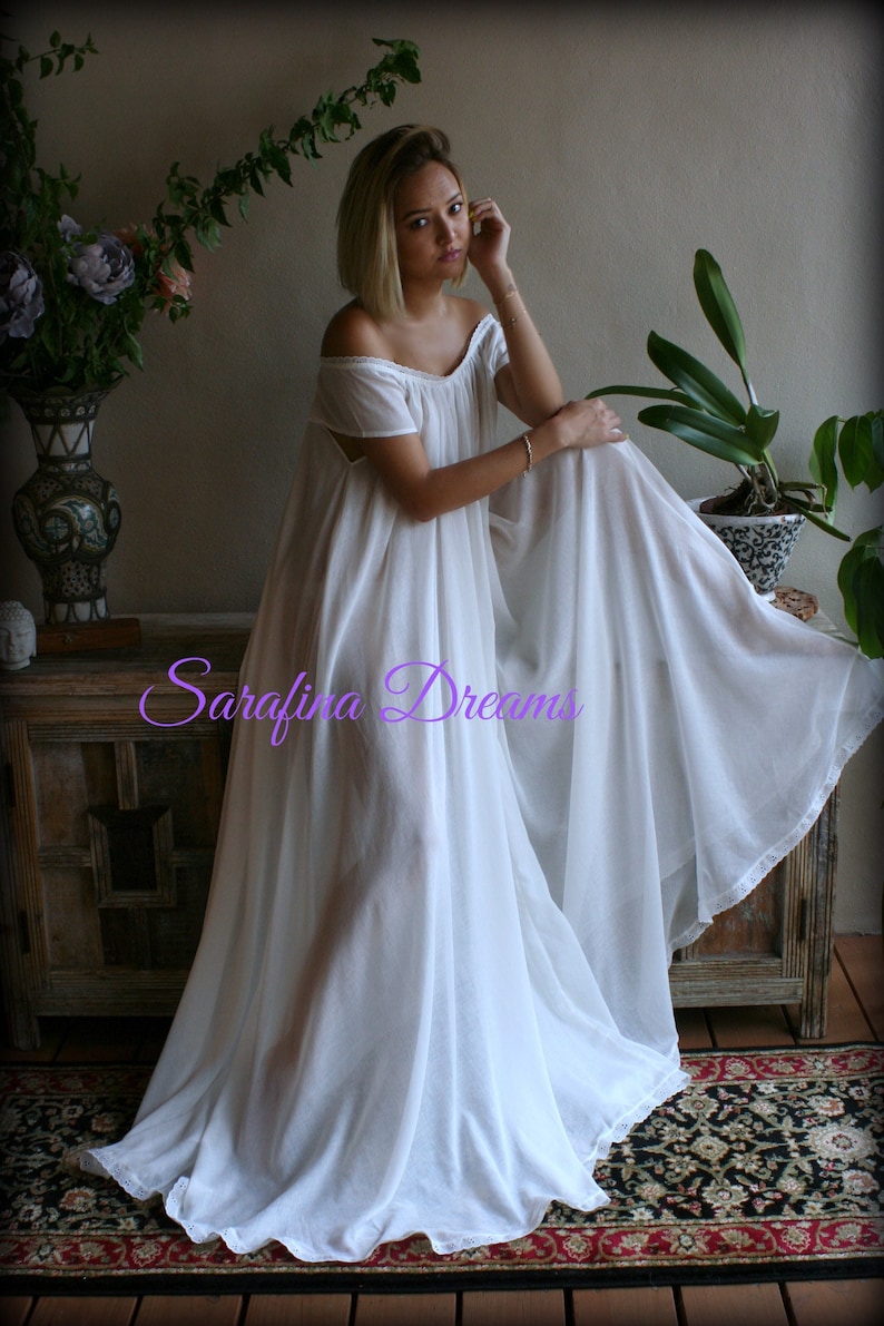 May include: A white, off-the-shoulder, long, flowing nightgown with a lace trim. The gown is made of a sheer fabric and has a relaxed fit. The model is wearing the gown and sitting on a wooden chest with a floral pattern. The text "Sarafina Dreams" is visible on the gown.