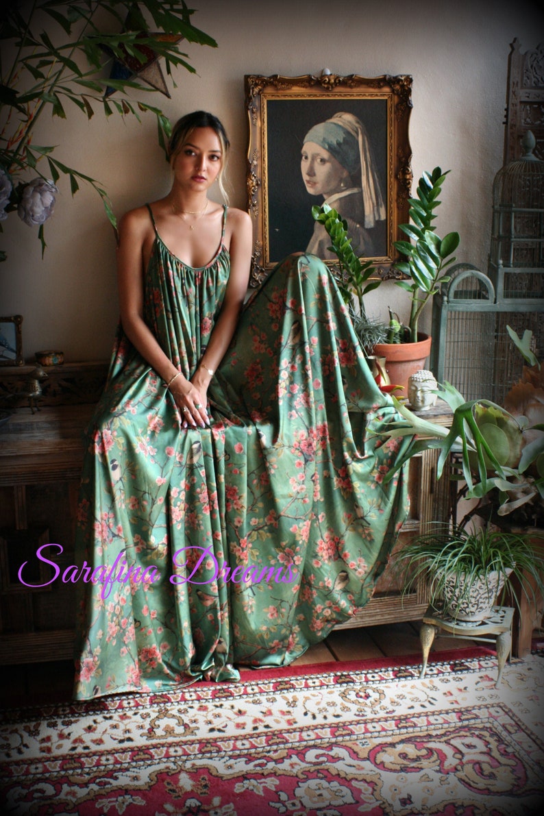 Puede incluir: Un vestido largo verde con un estampado floral de flores rosas y p&aacute;jaros. El vestido tiene tirantes finos y un escote fruncido. La imagen tambi&eacute;n presenta una pintura, plantas y una jaula decorativa para p&aacute;jaros.