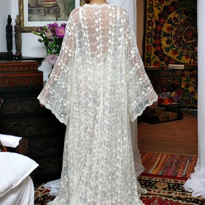 Puede incluir: Un vestido largo de encaje color crema con un estampado floral. El vestido tiene mangas largas y una falda fluida. La persona est&aacute; de pie en una habitaci&oacute;n con una alfombra roja estampada y una pintura en la pared. El vestido es una pieza delicada y elegante.