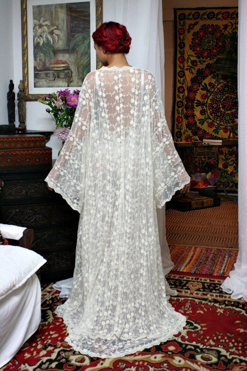 Puede incluir: Un vestido largo de encaje color crema con un estampado floral. El vestido tiene mangas largas y una falda fluida. La persona est&aacute; de pie en una habitaci&oacute;n con una alfombra roja estampada y una pintura en la pared. El vestido es una pieza delicada y elegante.